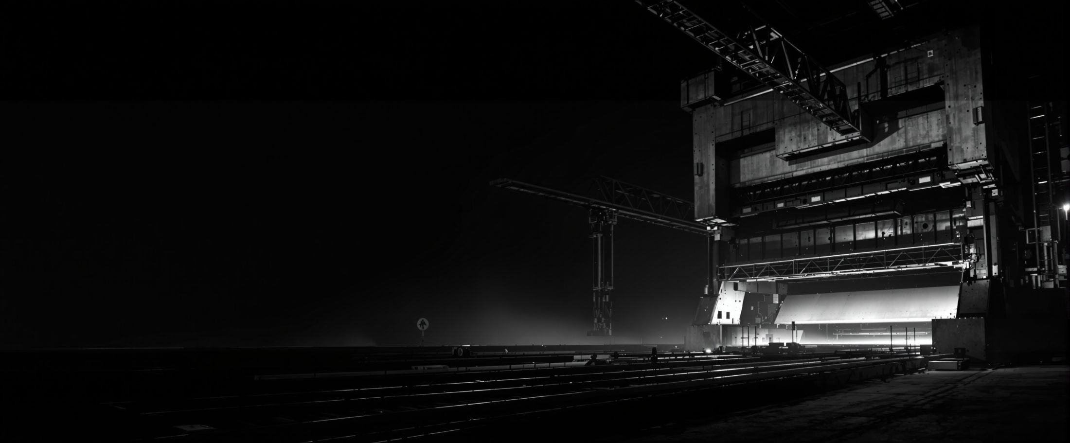 Planta de maquinaria pesada de noche: una prensa de acero y el raíl de un puente
        grúa iluminados por luz de trabajo dura, con la nave a oscuras alrededor.
